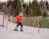 Dziecko w czerwonej kurtce i kasku narciarskim na stoku, obok siatki zabezpieczającej.