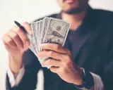 Mężczyzna w garniturze trzyma plik banknotów 100 USD. To wizualizacja rachunku przepływów pieniężnych krok po kroku.