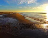 Två personer promenerar längs stranden vid Skagens udde i solnedgången. Vågorna sköljer in mot land.