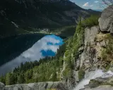 Gdzie jest Morskie Oko? Jezioro odbija chmury, otoczone lasem i górami. Wodospad spada po skalnej ścianie.