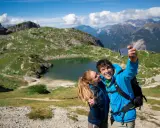 Coppia felice si scatta un selfie con il lago Coldai sullo sfondo, circondato da montagne verdi e un cielo azzurro con nuvole bianche.