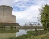Kühltürme eines Atomkraftwerks in Frankreich, die Dampf abgeben. Ein ruhiger See spiegelt die Anlage wider, während am Ufer Bäume und Gras wachsen.