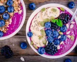 Zwei Schalen mit gesunden Bowls, gefüllt mit lila Smoothie, garniert mit Blaubeeren, Bananenscheiben, Brombeeren, Kürbiskernen und Granola.