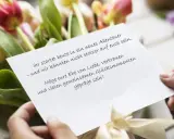 Hände halten eine Karte mit Glückwünschen vor einem Blumenstrauß. "Alles liebe zu eurem Hochzeitstag!" steht darauf.
