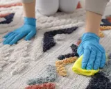Ręce w niebieskich rękawiczkach czyszczą kolorowy dywan. Jedna ręka przesuwa się po włóknach, druga z żółtą ściereczką delikatnie wyciera. Dowiedz się, jak wyczyścić dywan na sucho.