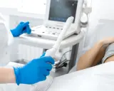 Medico con guanti blu tiene sonda per ecografia transvaginale. Paziente sdraiata sullo sfondo.