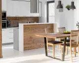 Cocina semiabierta al salón con barra de madera, encimeras blancas y electrodomésticos integrados. Mesa de comedor de madera con sillas.