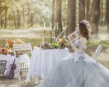 Kobieta w sukni ślubnej czyta książkę przy stole zastawionym owocami i szampanem. Sceneria leśna, atmosfera spokoju przed weselem.