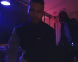 Dwóch mężczyzn w ciemnym pomieszczeniu, oświetlonym niebieskim i różowym światłem. Jeden z nich, 730 huncho, ma na sobie bluzę z logo Nike.