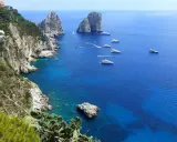 Scogliere verdi e rocce imponenti si affacciano sul mare blu intenso, con yacht che navigano. Un'immagine che ispira su come raggiungere Capri da Napoli.