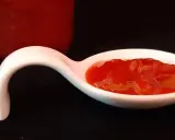 Une cuillère blanche présente une portion de confiture fraise rhubarbe, d'un rouge vif et appétissant. Un pot de confiture est visible en arrière-plan.