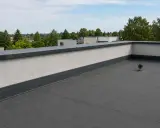Nowoczesna attyka na dachu z szarym obrzeżem i białym tynkiem, otaczająca płaski, pokryty papą dach.