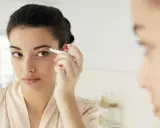 Une jeune femme ajuste ses sourcils broussailleux avec une pince à épiler devant un miroir, dans une tenue de chambre satinée.