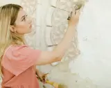 Młoda kobieta z piegami i blond włosami maluje pędzlem ozdobną sztukaterię na ścianie.