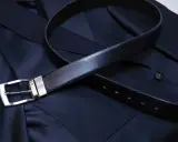 A black leather belt with a silver buckle rests on a dark navy suit jacket. Do you wear a belt with a tuxedo? This image shows a classic accessory.