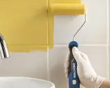 Mano con guante blanco pintando azulejos de cocina de blanco a amarillo con rodillo.