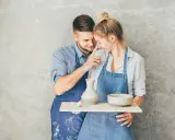 Para świętuje 9 rocznicę ślubu, tworząc ceramikę. Mężczyzna delikatnie dotyka nosa kobiety, oboje ubrani w fartuchy, ubrudzeni gliną.