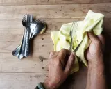 Mains polissant des couverts en argent avec un chiffon jaune. Un tas de fourchettes et cuillères est à gauche. Apprenez comment nettoyer l'argenterie.
