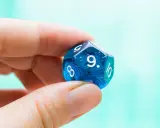 Palec i kciuk trzymają niebieską, dwudziestościenną kostkę z błyszczącymi drobinkami. Widoczna jest cyfra 9, symbolizująca numerologiczną 9.