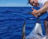 Uomo felice mostra il suo **biggest tuna** appena pescato in mare aperto, con una barca e una vela all'orizzonte.