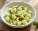Salade melon feta rafraîchissante, parsemée de menthe fraîche et de graines de sésame, servie dans un bol blanc sur une table en bois.
