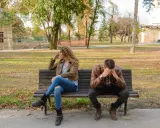 Pareja sentada en un banco, ella mira a un lado, él se tapa la cara. Parecen estar intentando procesar amor y desamor en un parque otoñal.