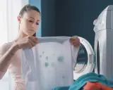Zmartwiona kobieta trzyma białą koszulkę z zielonymi plamami, która wygląda jak zafarbowane ubrania. Obok stoi otwarta pralka.