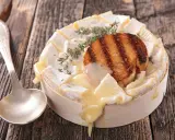 Un camembert coulant, plat typique normandie, garni d'une tranche de pain grillé et de thym, prêt à être dégusté avec une cuillère.
