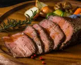 Succulento arrosto di manzo affettato, servito su un tagliere di legno con verdure arrosto, cipolle e rosmarino.