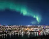 Grön norrskensbåge lyser upp natthimlen över en stad vid vattnet. En vacker demonstration av hur bildas norrsken.