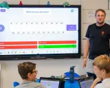 Un insegnante guida una lezione interattiva, simile a Kahoot, con domande su uno schermo touch. I bambini partecipano con entusiasmo.