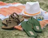 Brown sandals vs. green flip flops on a beach towel, with a hat and body mist. A perfect beach day awaits!