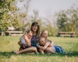 Mama z córkami na trawie, uśmiechnięte i przytulone. Widać, że czują motyle w brzuchu od tej miłości.