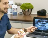 Dentysta omawia z pacjentką plan leczenia, pokazując na ekranie zdjęcie rentgenowskie szczęki i model zębów, wyjaśniając znaczenie czyszczenia dziąseł.