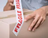 Ręce zaklejają karton taśmą z napisem "FRAGILE". Uwaga szkło, kurier musi być ostrożny.