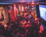 Un locale affollato a Barcellona by night, con uomini che socializzano e bevono. L'atmosfera è vibrante, illuminata da luci rosse e blu.
