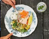 En tallrik med svensk lax, potatis, grönsaker och en citronskiva. En person skär i laxen med en gaffel och kniv.
