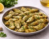 Carciofi in padella, teneri e dorati, conditi con erbe aromatiche e aglio. Accanto, spicchi d'aglio, prezzemolo fresco e una fetta di limone.
