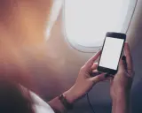 Kobieta trzyma telefon przy oknie w samolocie. Czy w samolocie można korzystać z telefonu? Tak, ale z ograniczeniami.