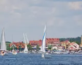 Piękne mazury widoki: żaglówki na jeziorze, miasteczko z czerwonymi dachami i wieżą kościelną w tle.