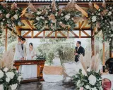 Novios celebran aniversario en altar decorado con flores y pampas. Un oficiante habla mientras la pareja sonríe.