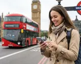 Kobieta z telefonem w ręku, na tle czerwonego autobusu i Big Bena. Wygodny internet w UK ułatwia zwiedzanie.