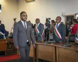 Des hommes en costume, certains portant des écharpes aux couleurs nationales, se tiennent dans une salle. L'un d'eux semble être une persona non grata, marchant à l'écart.