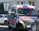 Ambulans z ratownikami medycznymi na tle budynku i białego samochodu.