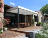 Terrasse en bois sous un toit pergola naturel, avec table et chaises, entourée de végétation luxuriante et d'une maison en pierre.