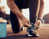 Uomo si allaccia le scarpe da corsa, pronto per correre a stomaco vuoto. Accanto, una borraccia blu.