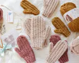 Mitones tejidos a mano con patrón de dos agujas. Varios pares en tonos rosa, beige y mostaza, junto a ovillos de lana y agujas.