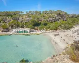 Türkisfarbenes Wasser und ein sonniger Sandstrand in Cala Pi, umgeben von grünen Hügeln. Menschen entspannen sich unter Sonnenschirmen.