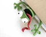 Marcador de libro con ranas amigurumi, verde y negro, con lengua roja, asomando de un libro beige.