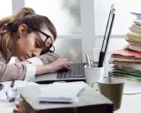 Zmęczona kobieta śpi na laptopie, otoczona stertami dokumentów. To obraz pracoholizmu, który doprowadził ją do wyczerpania.